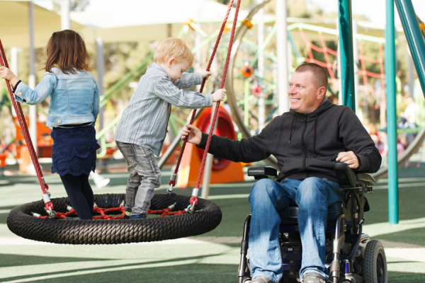 A father dressed in a dark sweatshirt and jeans is seated in a wheelchair plays with his two young children on a tire swing at a playground